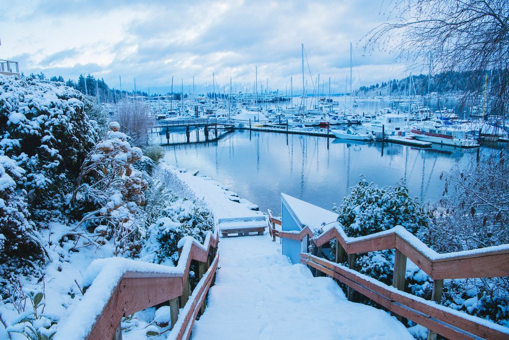 Snow-covered wooden stairs lead down to a marina with multiple boats docked under a cloudy winter sky in Washington, USA.