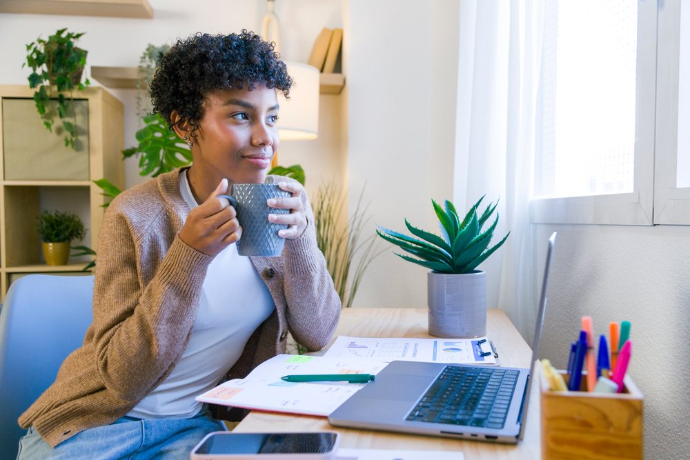 Woman drinking coffee at her desk with laptop and notebook in a home office under natural light, remote work.