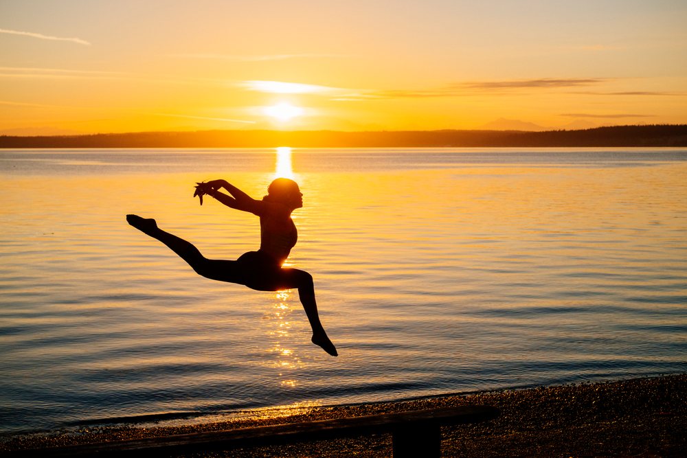 Silhouette of a woman leaping in a yoga or acrobatic pose by a lake at sunset, USA.