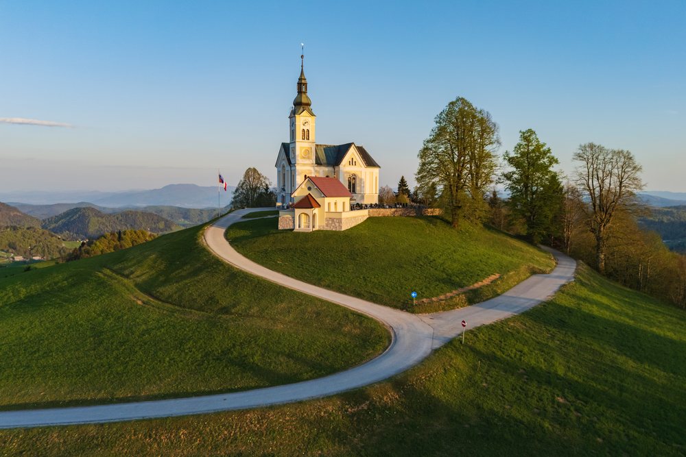 Parish church of St Leonards sits atop a green hill with curved paved roads, leafy trees, and a Slovenian flag, landscape, cityscape, Polhov Gradec, Slovenia.