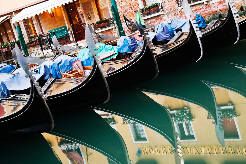 Six black gondolas with blue covers docked side by side in a narrow Venetian canal reflecting historic brick and plaster buildings with flowered windows, Veneto, Italy.