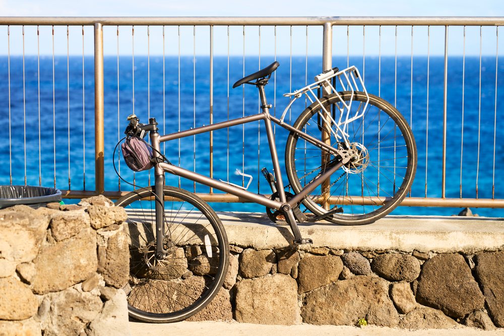 Bicycle with black saddle leaning against metal railing and stone wall by clear ocean water in sunlight, Oahu, Hawaii, USA.
