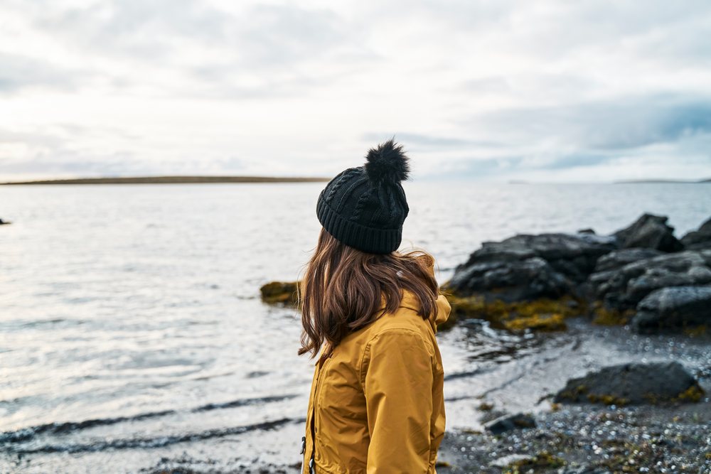 White Woman stands on rocky shoreline looking out over calm water