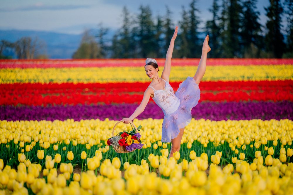 Ballet dancer in costume and crown holding a flower basket, standing in a colorful tulip field under clear blue sky, USA.