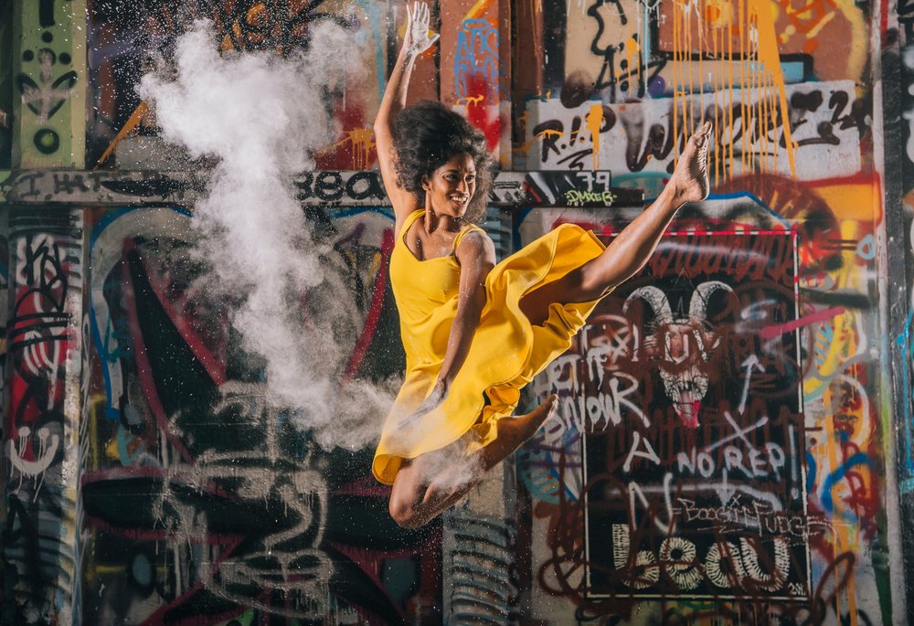Woman in a yellow dress jumps joyfully throwing powder before a colorful graffiti wall, USA.