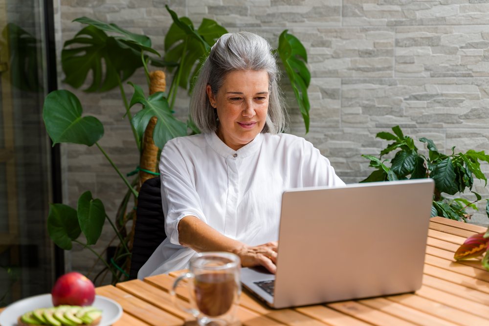 Woman with gray hair working on a laptop at a wooden table with coffee and a plate of fruit indoors, surrounded by plants, Spain.
