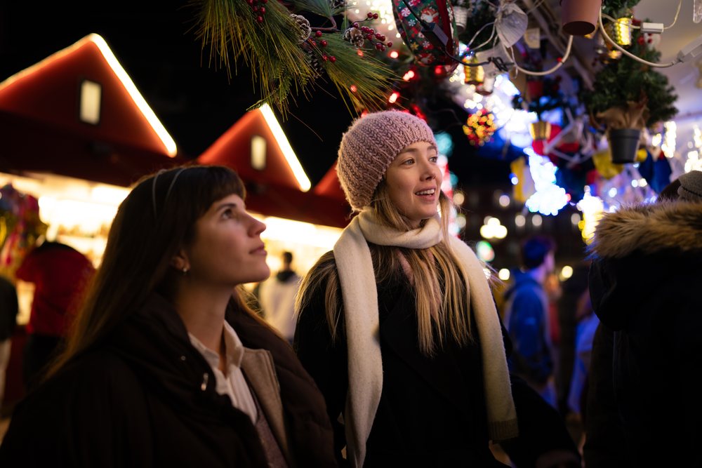 Two women wearing winter hats and scarves shop at a brightly lit Christmas market decorated with pine branches, red berries, and balls in Madrid at night, Spain.
