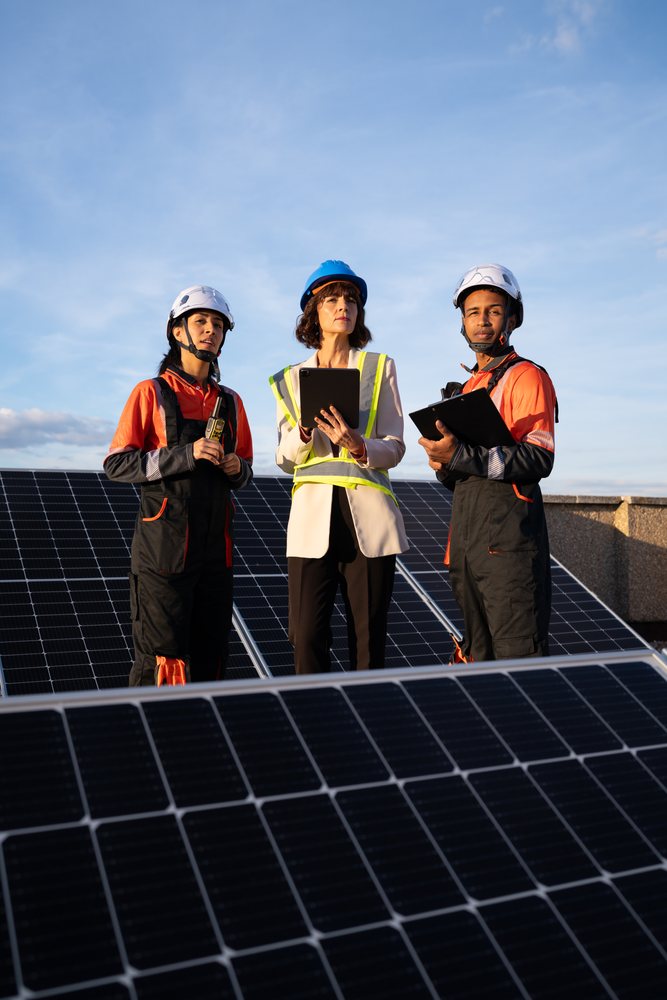 Three engineers wearing helmets and safety vests inspect solar panels outdoors under a clear sky, Madrid, Spain.