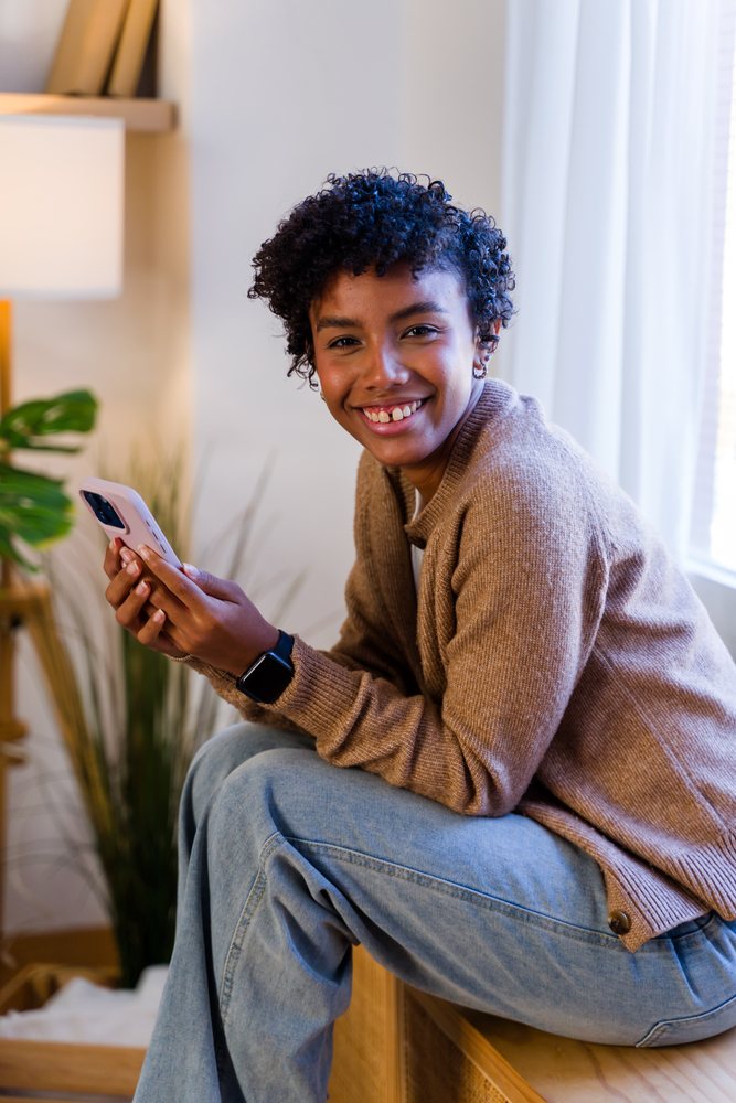 Woman indoors smiling while sitting and holding a smartphone by a window with plants and wooden furniture, Spain.