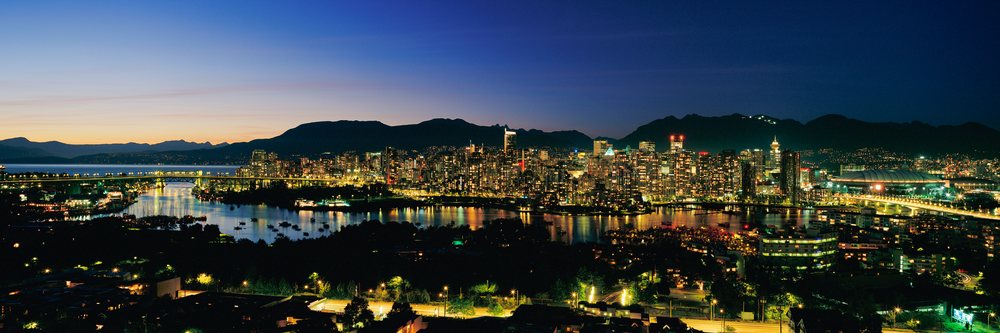 Illuminated Vancouver skyline at night with high-rise buildings, bridge over water, and mountains beyond.