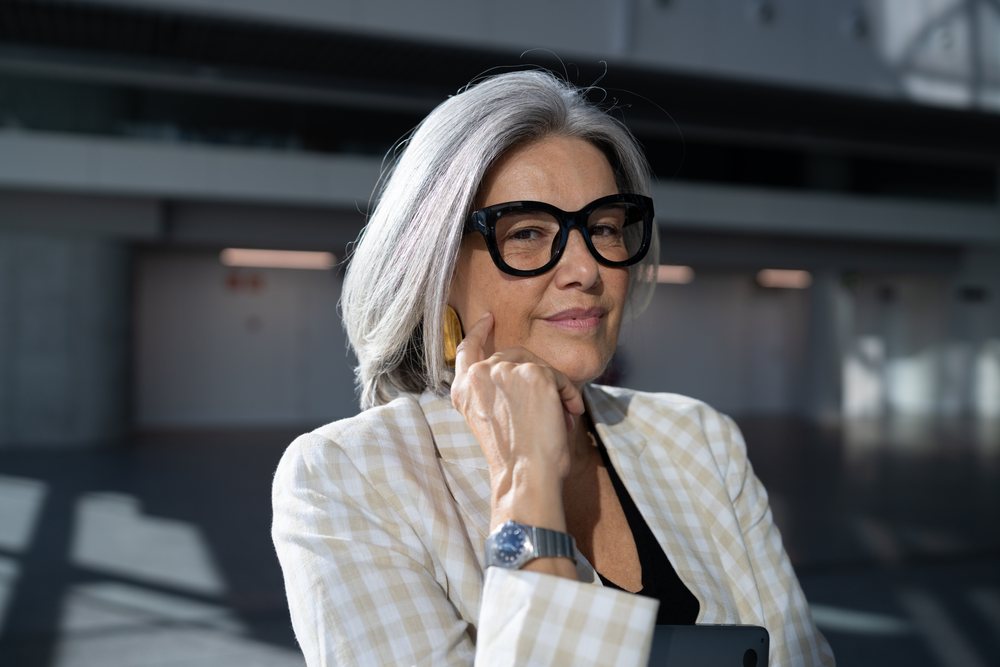 Mature woman with gray hair and black glasses wearing a plaid blazer indoors in a thoughtful pose, businesswoman, Spain, Madrid.
