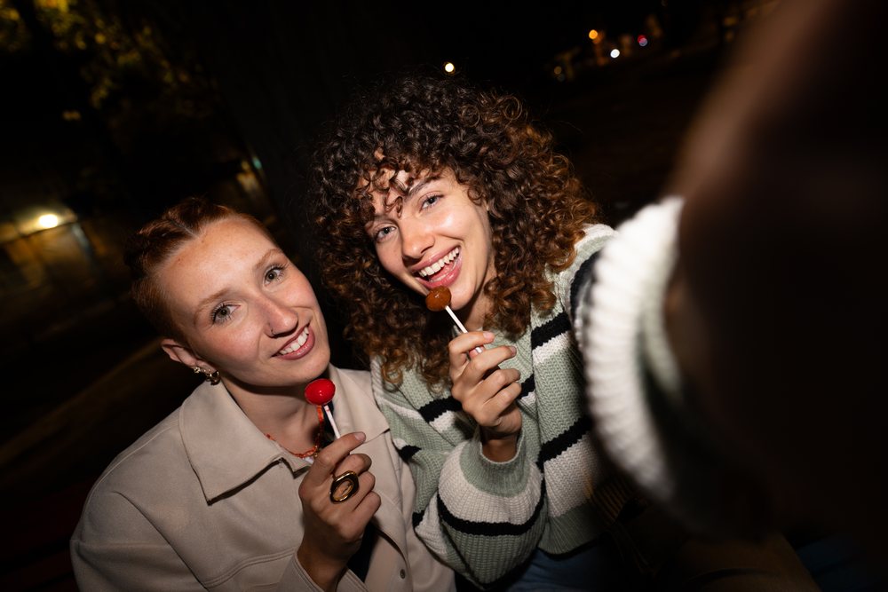 Two women with curly and straight hair smiling and holding lollipops while taking a selfie outdoors at night, friendship, Santiago De Compostela, Spain.