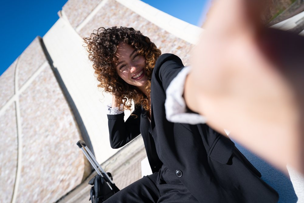 Businesswoman in black suit smiling and taking selfie outdoors near a modern stone building in Santiago de Compostela, traveling executive, Spain, Antiago De Compostela.