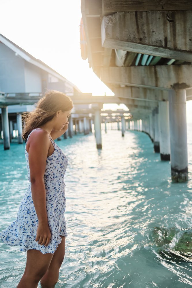 A young woman in a blue floral dress walks in the clear ocean water under a pier in the Maldives at sunset.