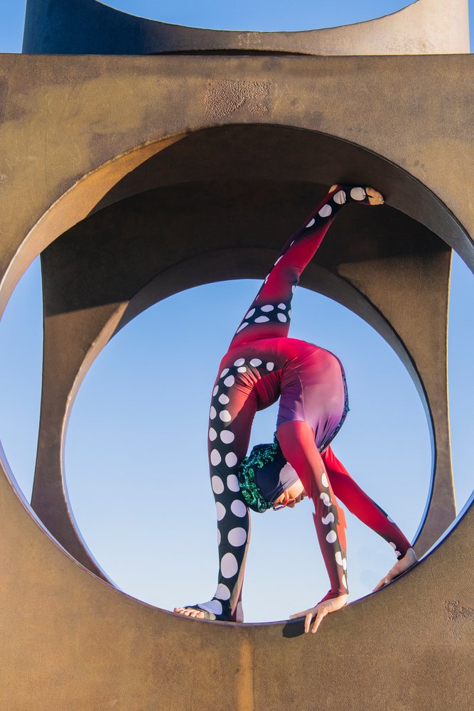 Contortionist in a red polka dot leotard balances in a circular arch outdoors under a clear blue sky, USA.