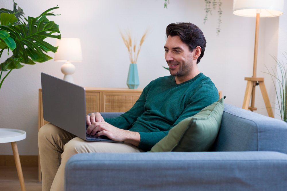 Man sitting on a sofa using a laptop in a modern living room decorated with plants and lamps, technology, young adult, home.