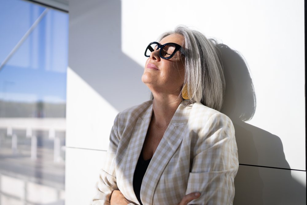 Mature woman with gray hair enjoys sunlight while leaning against a white wall, businesswoman, traveler, Spain, Madrid.