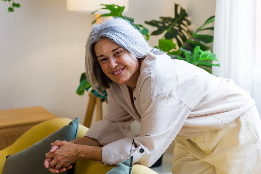 Mature woman with gray hair smiles while leaning on a yellow armchair with green cushions in a home interior, Spain.