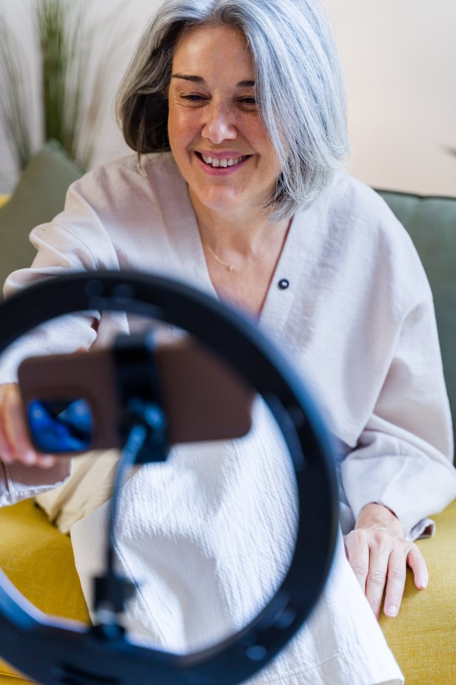 Smiling woman with gray hair holding a smartphone in front of a ring light indoors on a couch with a yellow cushion, Spain.