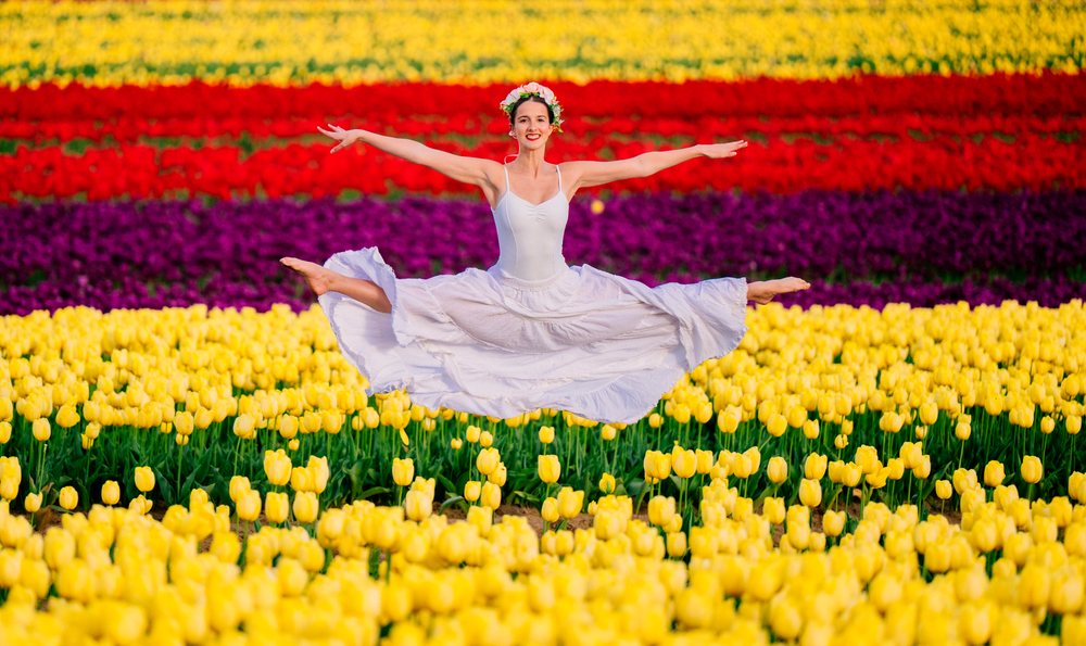 Ballet dancer in a white dress leaps gracefully wearing a flower crown in a colorful tulip field outdoors, USA.