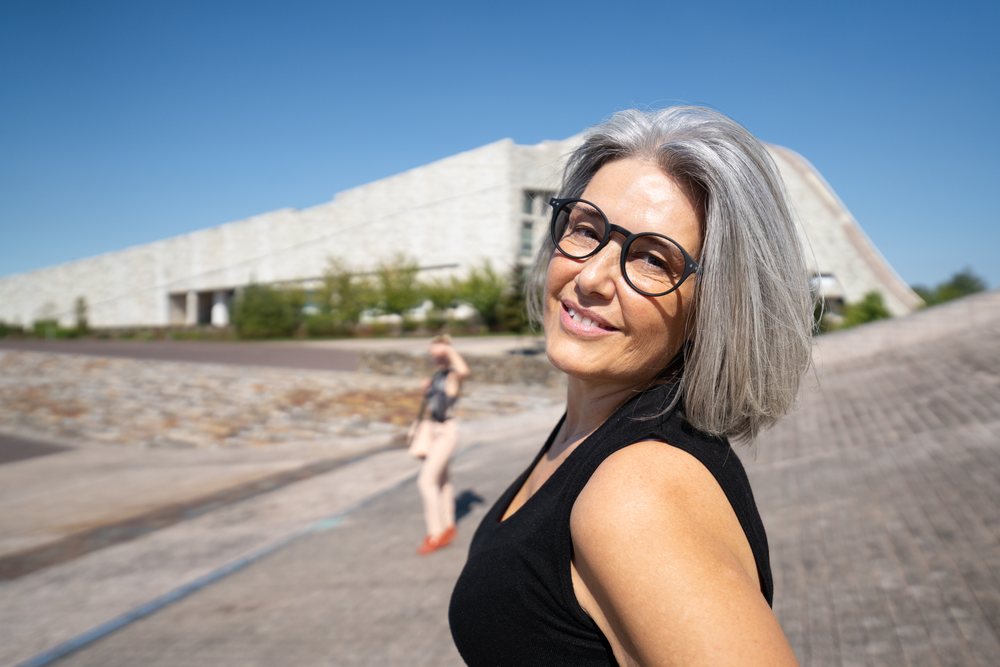 Smiling mature woman with gray hair and glasses stands outdoors near a modern building under a clear sky, businesswomen, Spain.
