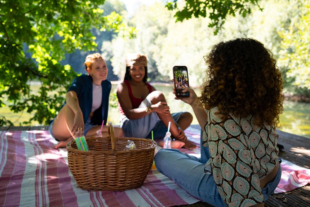 Three women sitting on a striped picnic blanket by a river, with one photographing the others using a smartphone outdoors, Spain.