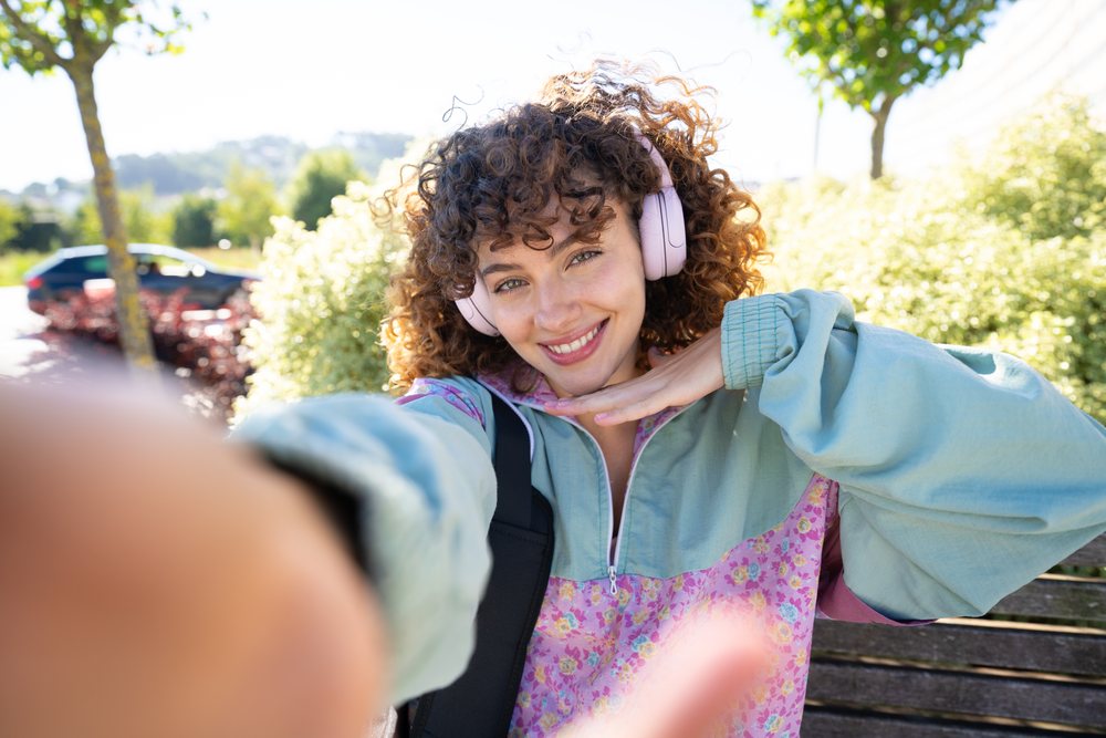 Woman with curly hair smiling and taking a selfie while sitting outdoors on a wooden park bench in spring or summer, Spain.