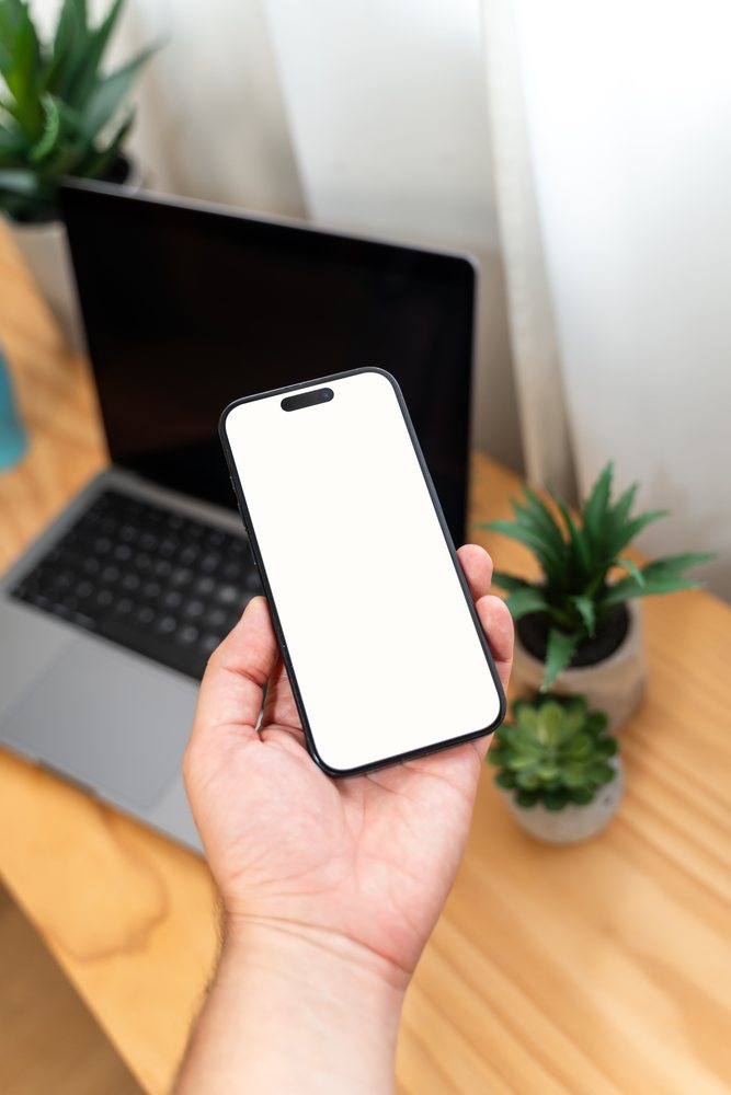 Hand holding a smartphone with a blank white screen above a wooden desk, mock up, Spain.