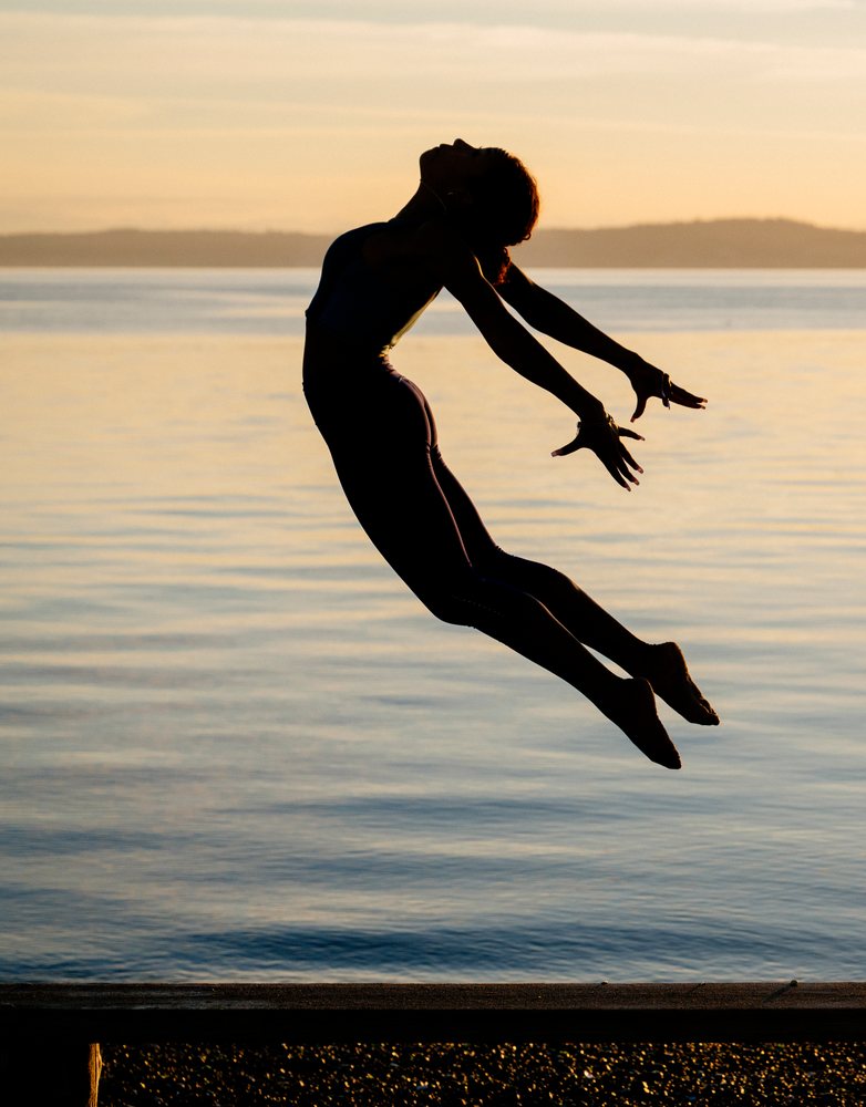 Woman jumping and posing in a yoga or acrobatic posture at sunset beside a lake, USA.