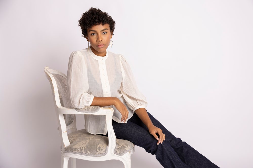 Young woman sits relaxed on a white antique wooden chair wearing a white buttoned blouse with puffed sleeves and dark denim jeans against a neutral plain background, fashion, young culture.