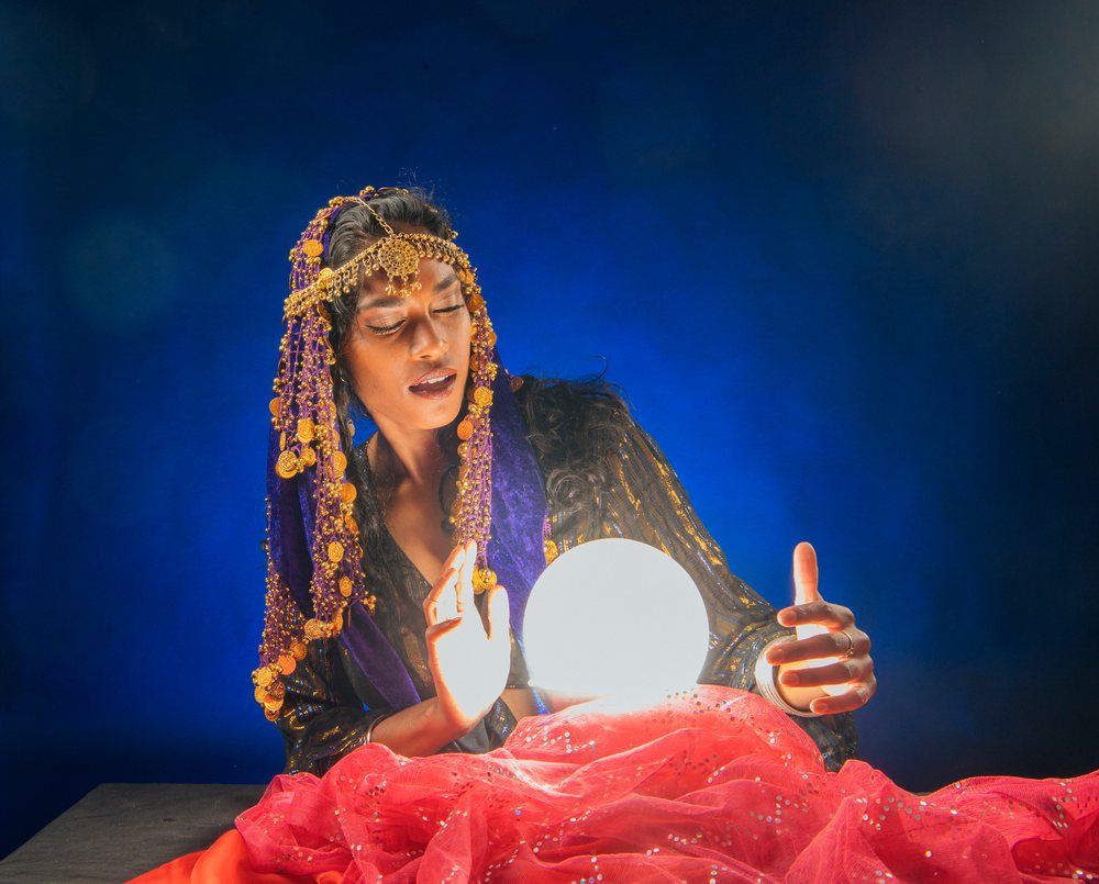 A fortune teller in traditional attire gazes into a glowing crystal ball as part of a spiritual divination practice, set against a dark background in the, USA.