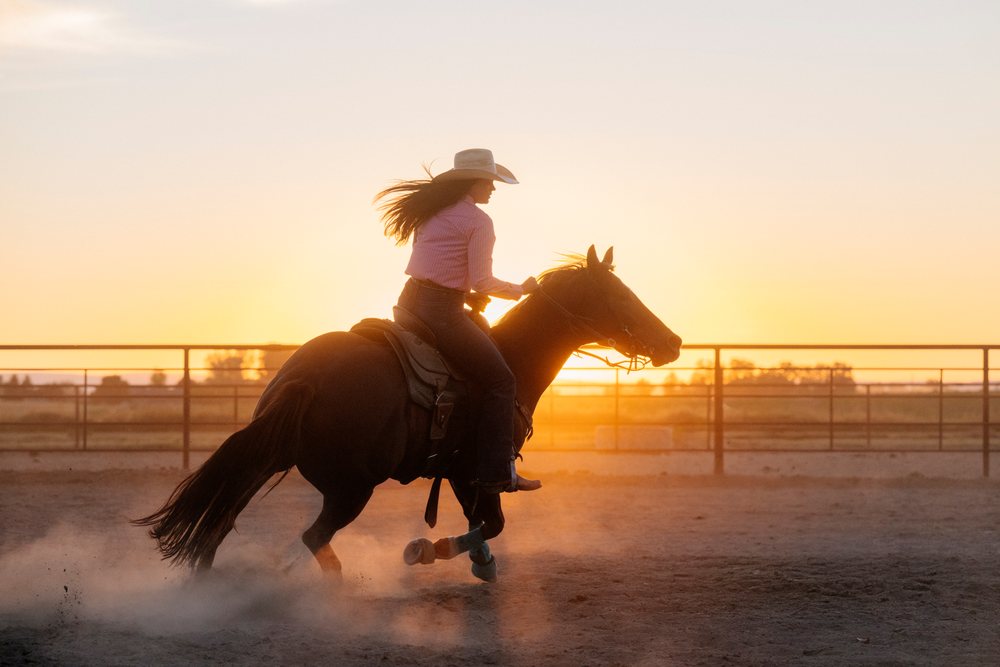 Woman wearing a cowboy hat and wrangler jeans rides a mare in a dusty corral at sunset on a rural Southwest USA ranch, professional.