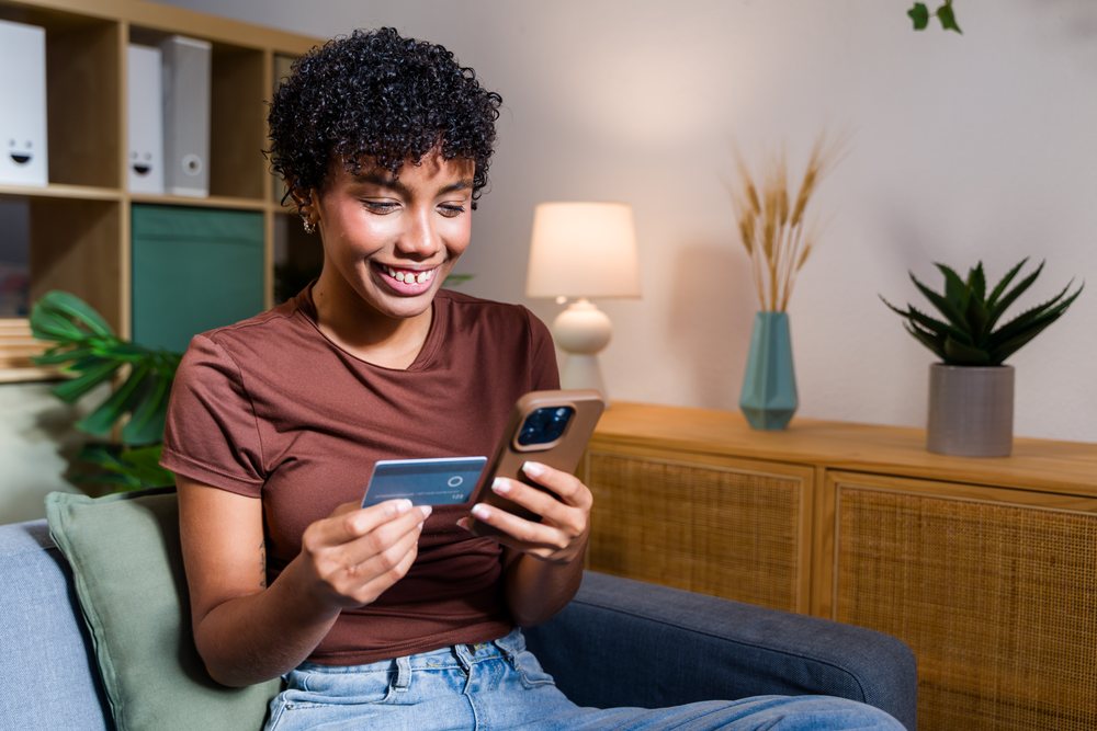 Woman with curly hair sits on a couch indoors holding a smartphone and credit card, smiling in a modern living room, Spain.