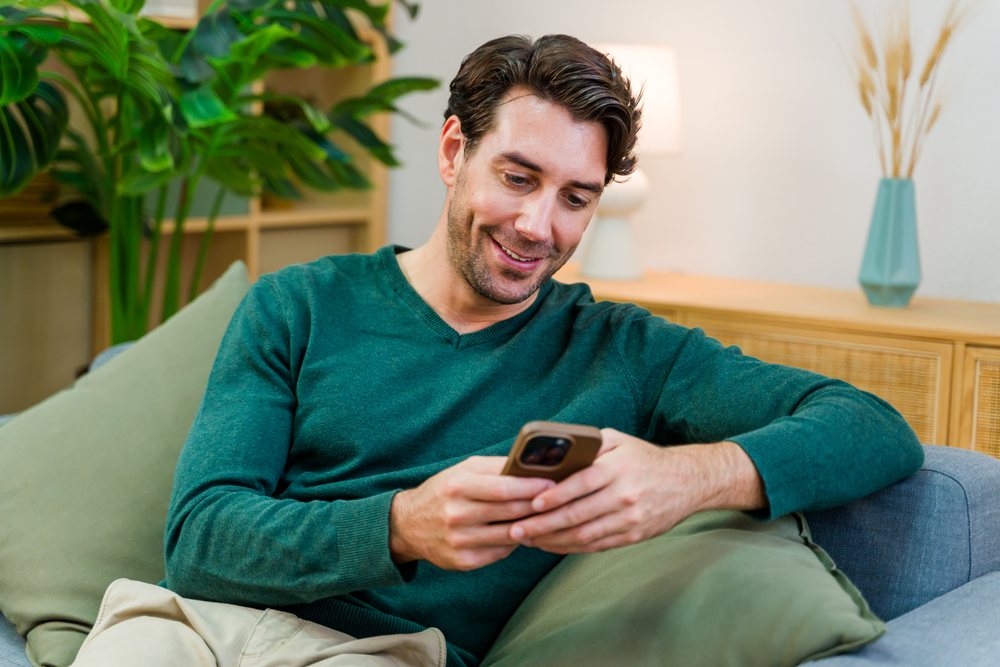 Man sitting on a couch smiling and using a smartphone in a living room with plants and decorative objects, Spain.
