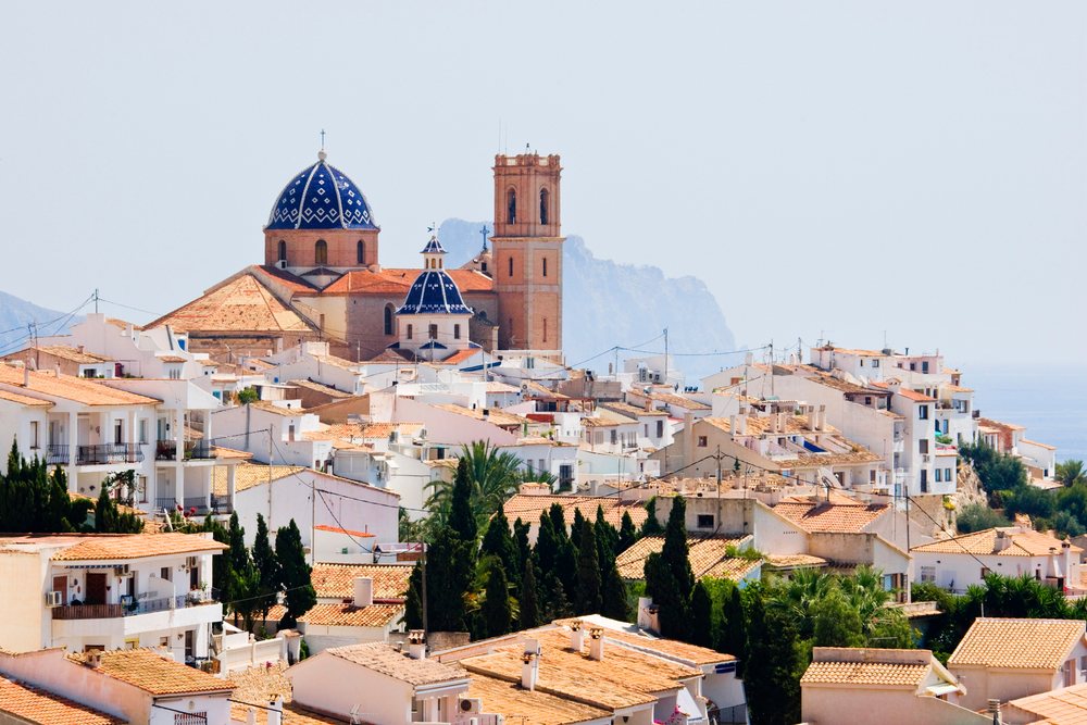 View of the Church of the Virgin del Consuelo with blue tiled domes and white terracotta-roofed buildings, with mountains and sea beyond, Costa Blanca, Spain.