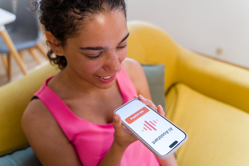 A woman sitting on a yellow sofa using an AI assistant application on her mobile phone at home, Spain.