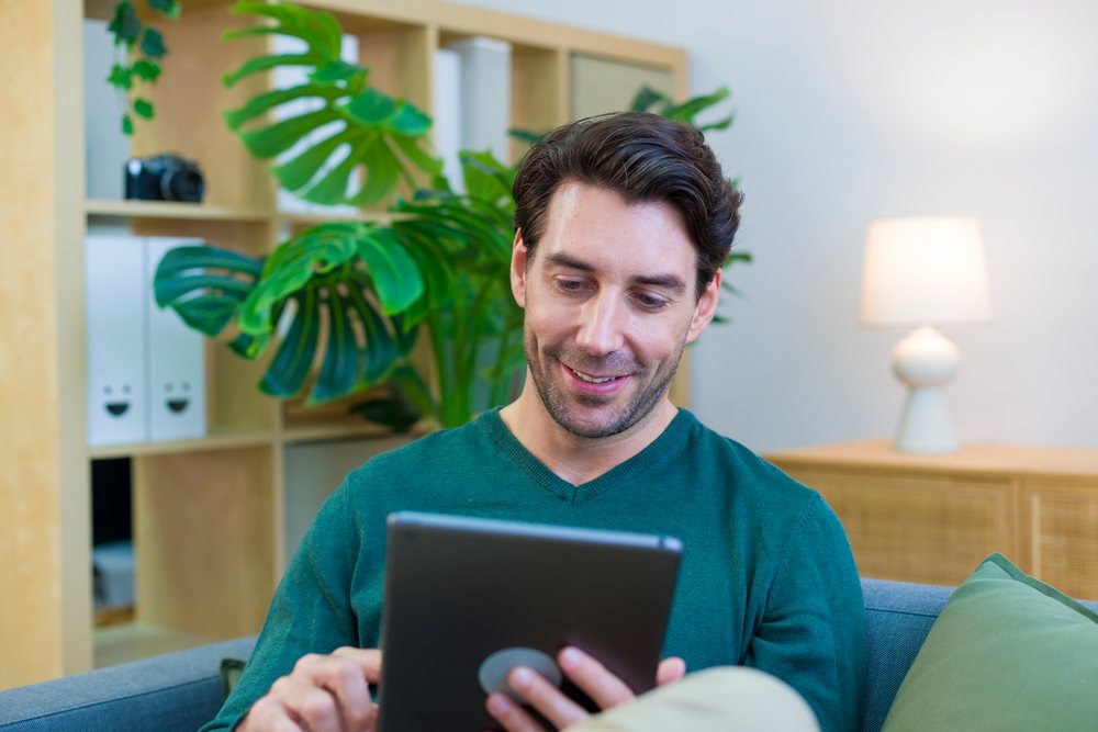 Young man sitting on a sofa using a tablet in a modern living room with houseplants and contemporary furniture, technology, young adult, home.