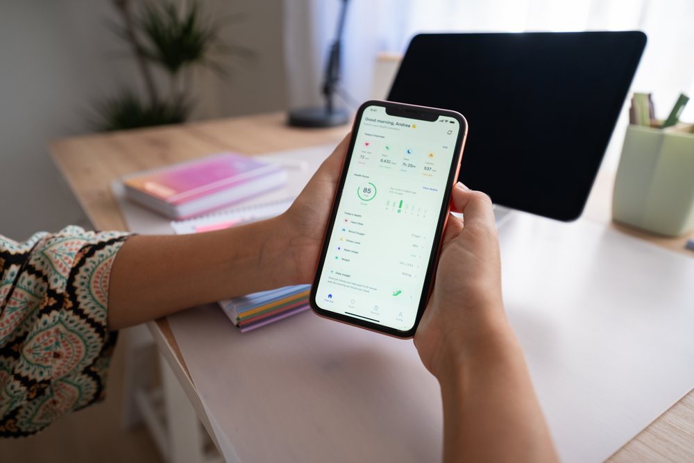 Person holding a smartphone displaying a health app while seated at a desk in a home office, Spain.