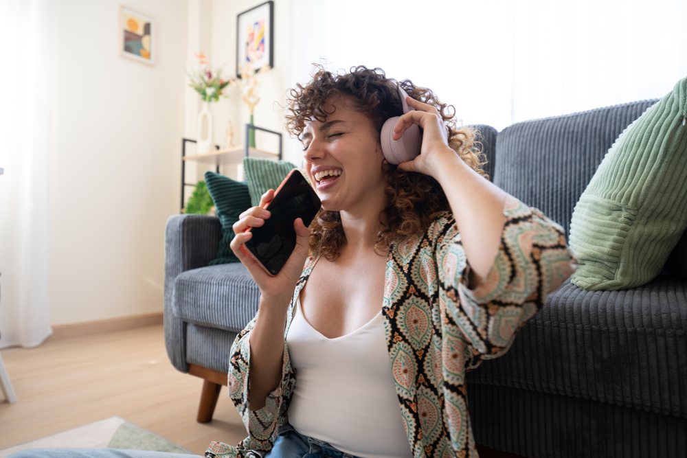 Woman sitting on floor in home living room in Spain wearing headphones and holding smartphone enjoying music.
