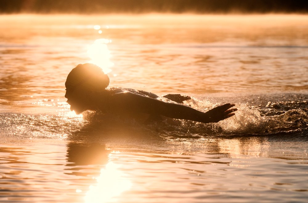 Person swimming in open water with sunlight reflecting off the surface during early morning exercise, USA.