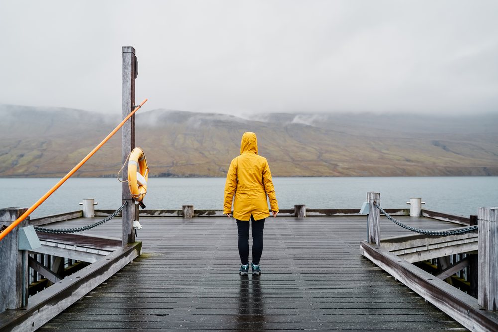 A person wearing a yellow raincoat stands on a wooden pier overlooking misty hills and water in Iceland under an overcast sky.