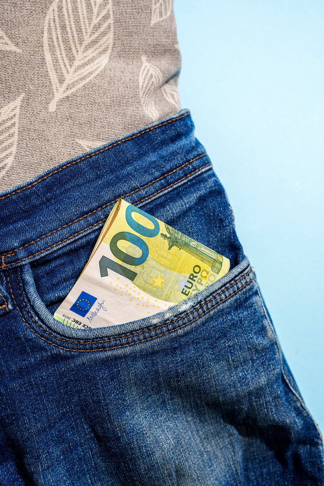 Rolled 100 euro banknote partially visible in a blue denim jeans pocket against a blue studio background, Studio Shot.