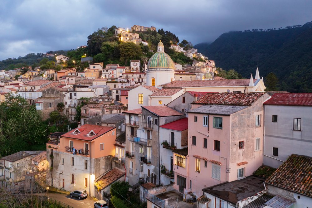 Sunset over the hill town of Nocera Terinese, showing an illuminated church dome and surrounding hillside buildings, Calabria, Italy.