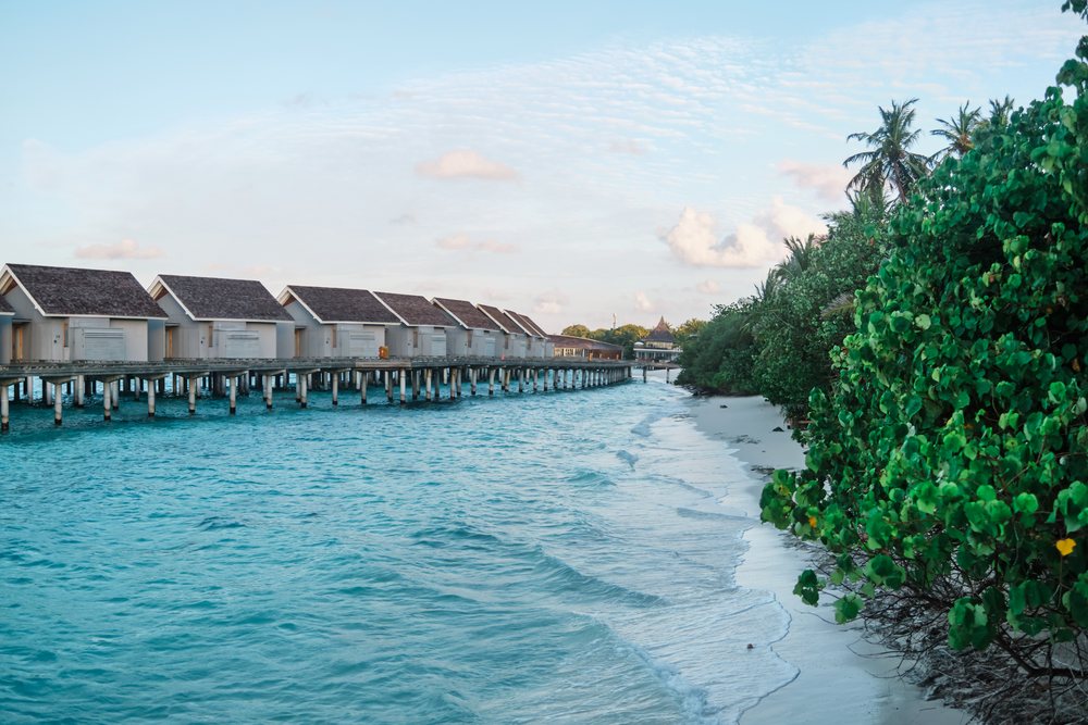 Water villas on stilts over turquoise ocean waters line a tropical beachfront resort alongside lush greenery and white sand in the, Maldives.