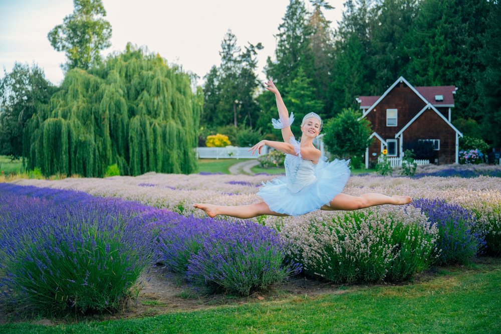 A ballerina in a white tutu performs a split leap gracefully outdoors in a lavender field surrounded by trees and greenery, WA, USA.