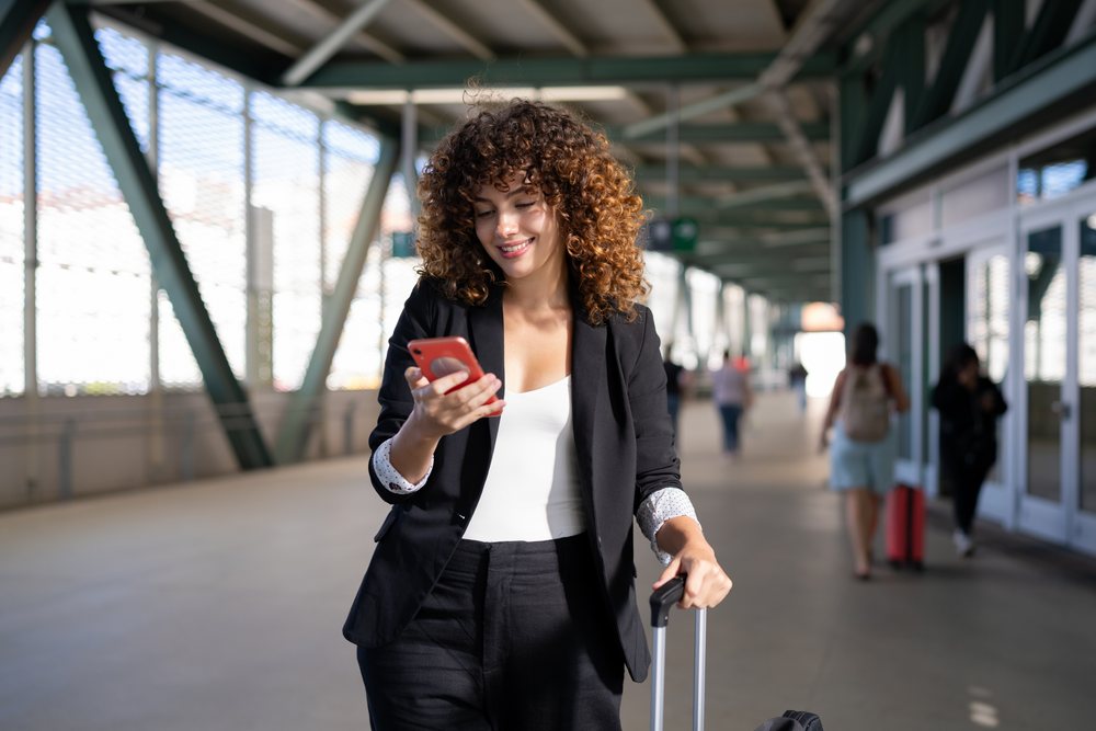 Woman with curly hair and black suit using smartphone while pulling rolling suitcase, Spain.