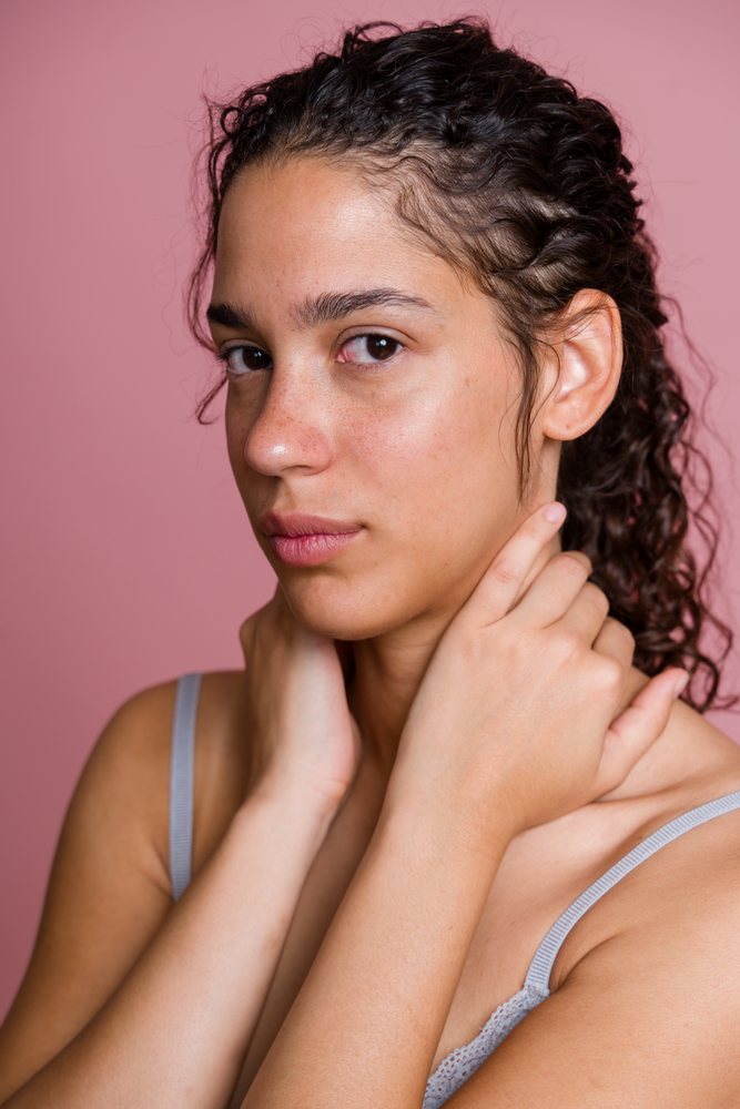 Studio portrait of a young woman with curly hair touching her neck against a pink background, beauty shoot, Spain.