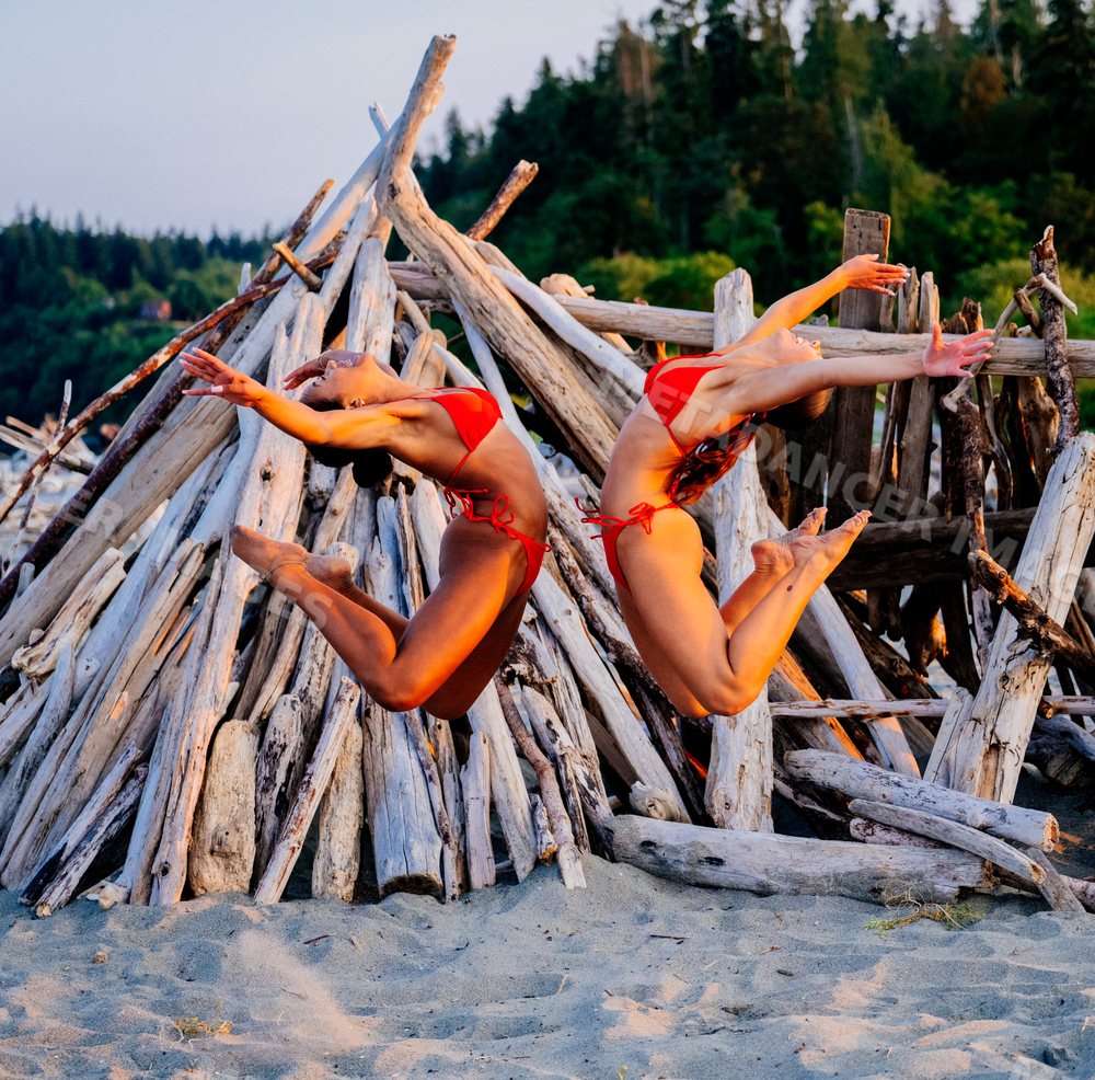 Two women jumping in red bikinis on a sandy beach with driftwood