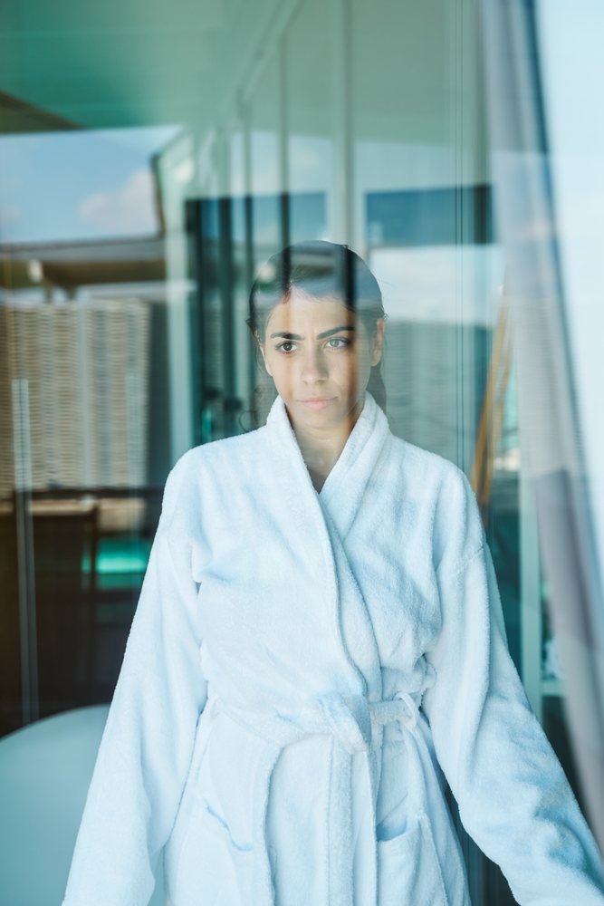 A woman wearing a white bathrobe looks pensively through a glass window inside a hotel room in the, Maldives.