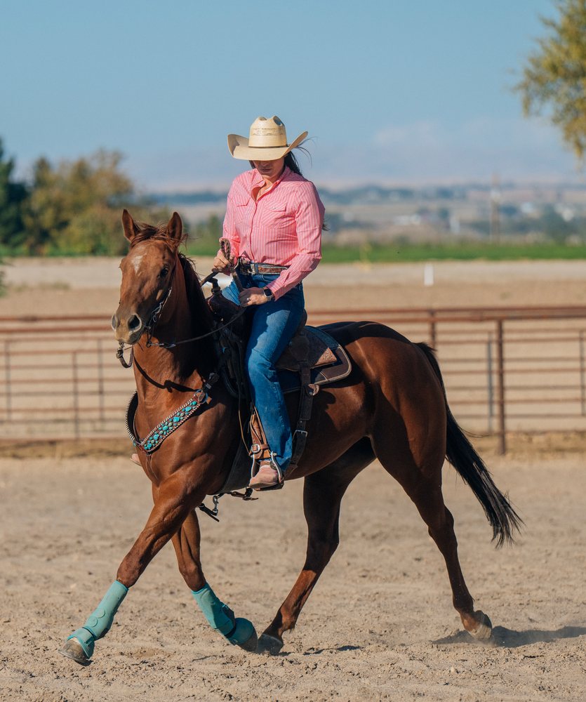 Woman wearing a cowboy hat rides a horse with turquoise leg wraps in a sandy outdoor arena, Southwest USA.